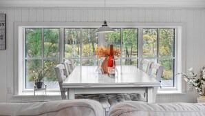 A dining area with a white table and chairs, a vase with flowers, and a view of trees through large windows. - 2 bedroom amazing home in Nexø (Nexø)