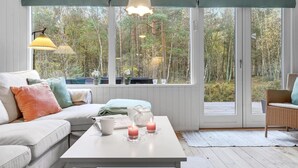 A cozy room with a white sofa, a coffee table with candles, and a view of a forest through large windows. - 2 bedroom amazing home in Nexø (Nexø)