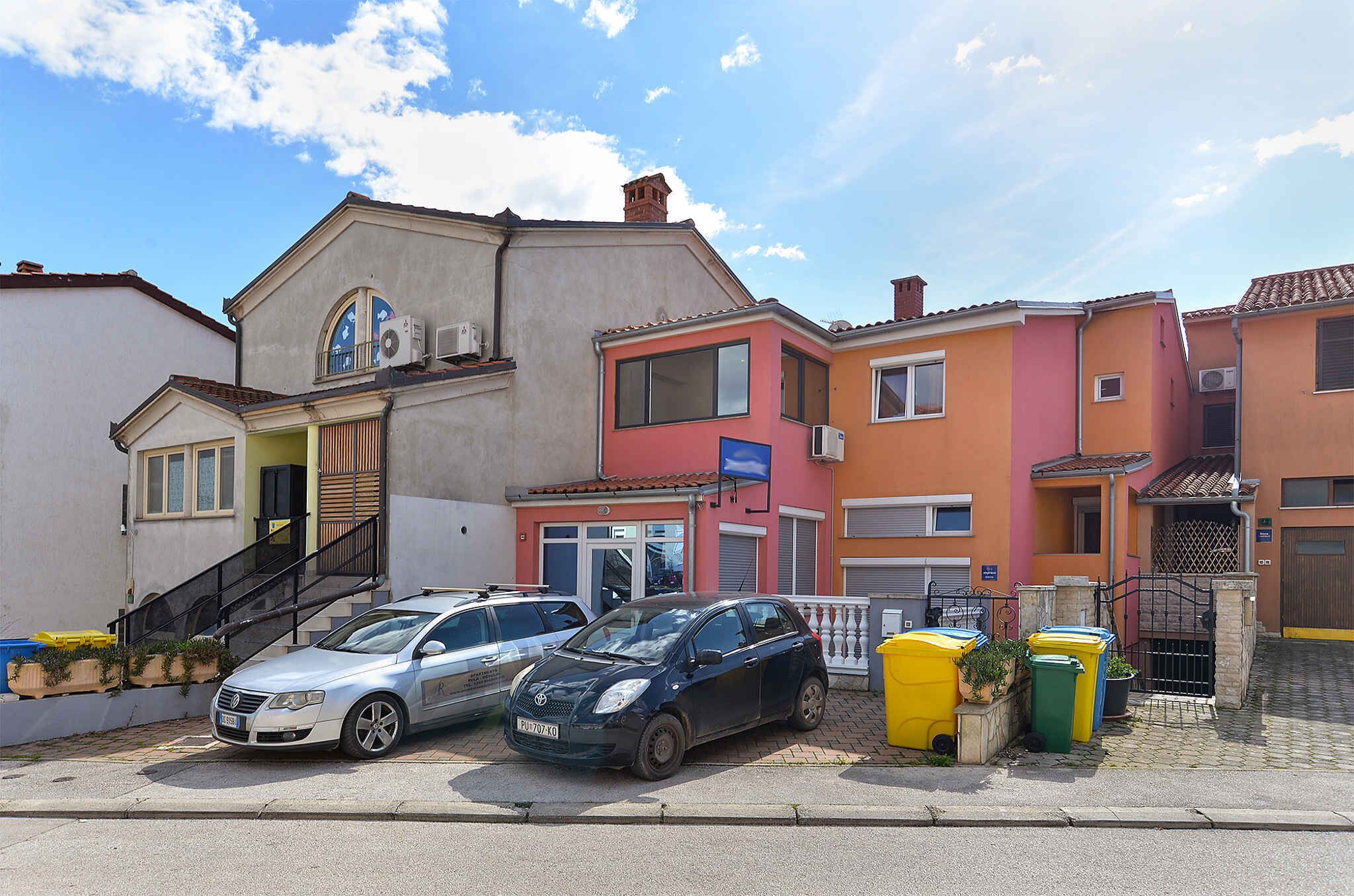 Une rangée de maisons aux couleurs vives, avec une voiture garée et des poubelles devant.