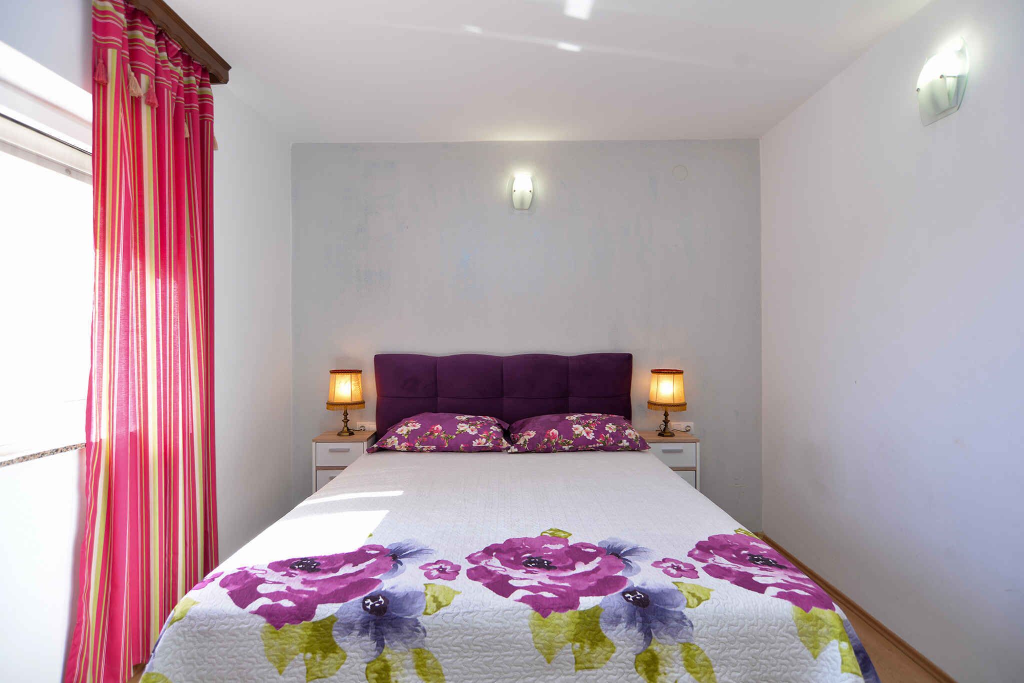 A bedroom with a bed, two bedside lamps, a headboard, and a window with curtains.
