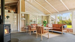 A cozy living room with a fireplace, wooden furniture, and large windows offering a view of the outdoors. - 3 bedroom beautiful home in Hjørring (Hjørring)