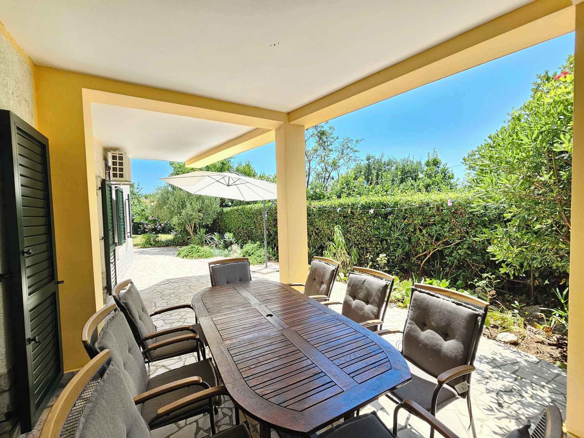 Eine Terrasse im Freien mit einem runden Tisch und sechs Stühlen, umgeben von gelben Wänden und einem überdachten Bereich.