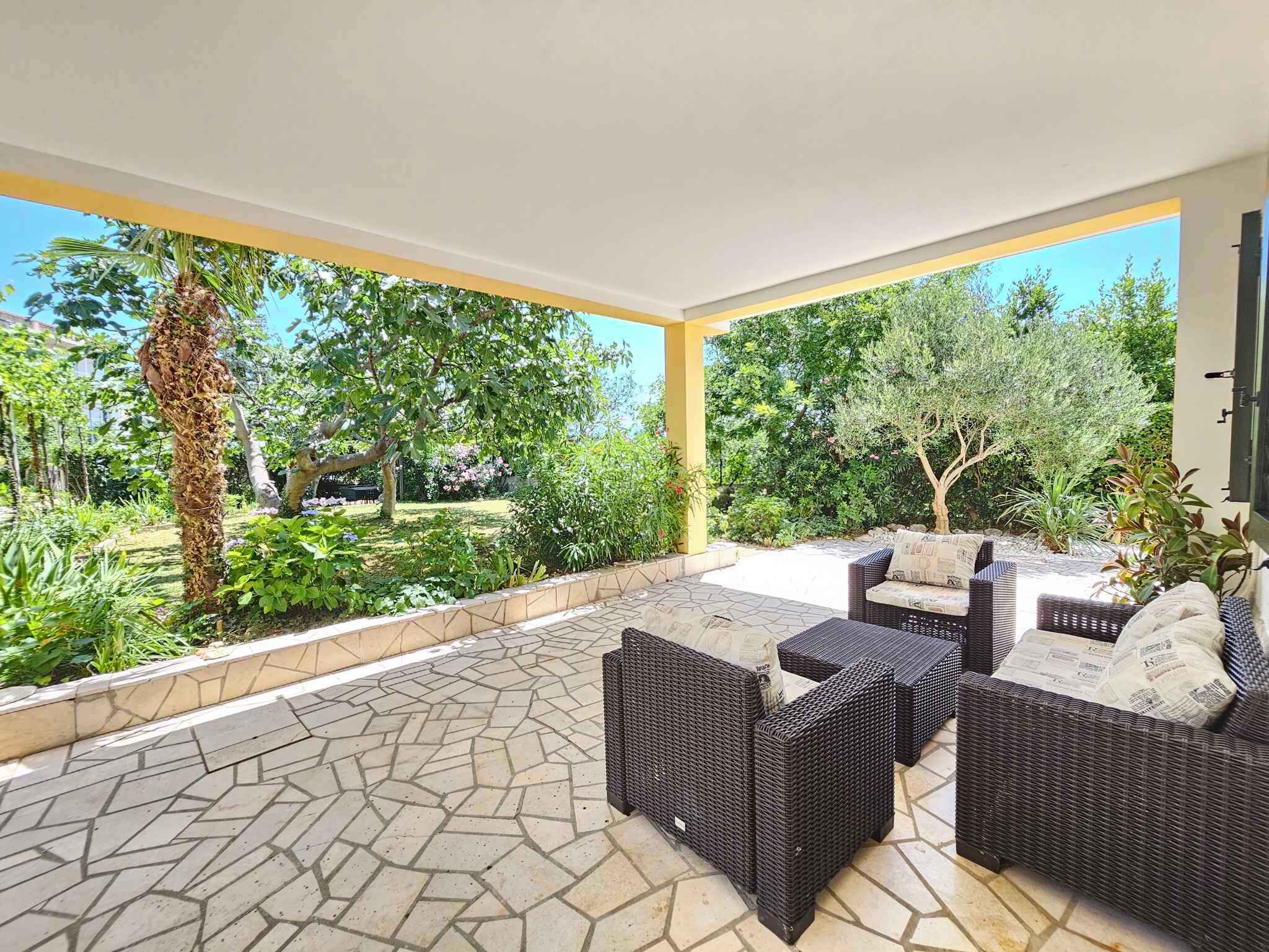 An outdoor patio with wicker furniture and a stone-tiled floor, surrounded by lush greenery and trees.