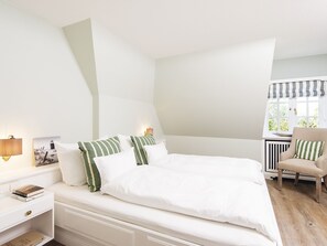 A bedroom with a large bed, white walls, a window with a view, a chair, and a nightstand with a lamp and a book.