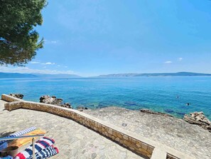 Terraza empedrada con vistas al mar azul y tranquilo, con rocas a la vista, un banco de madera y una tela a rayas cubriendo parte de él.