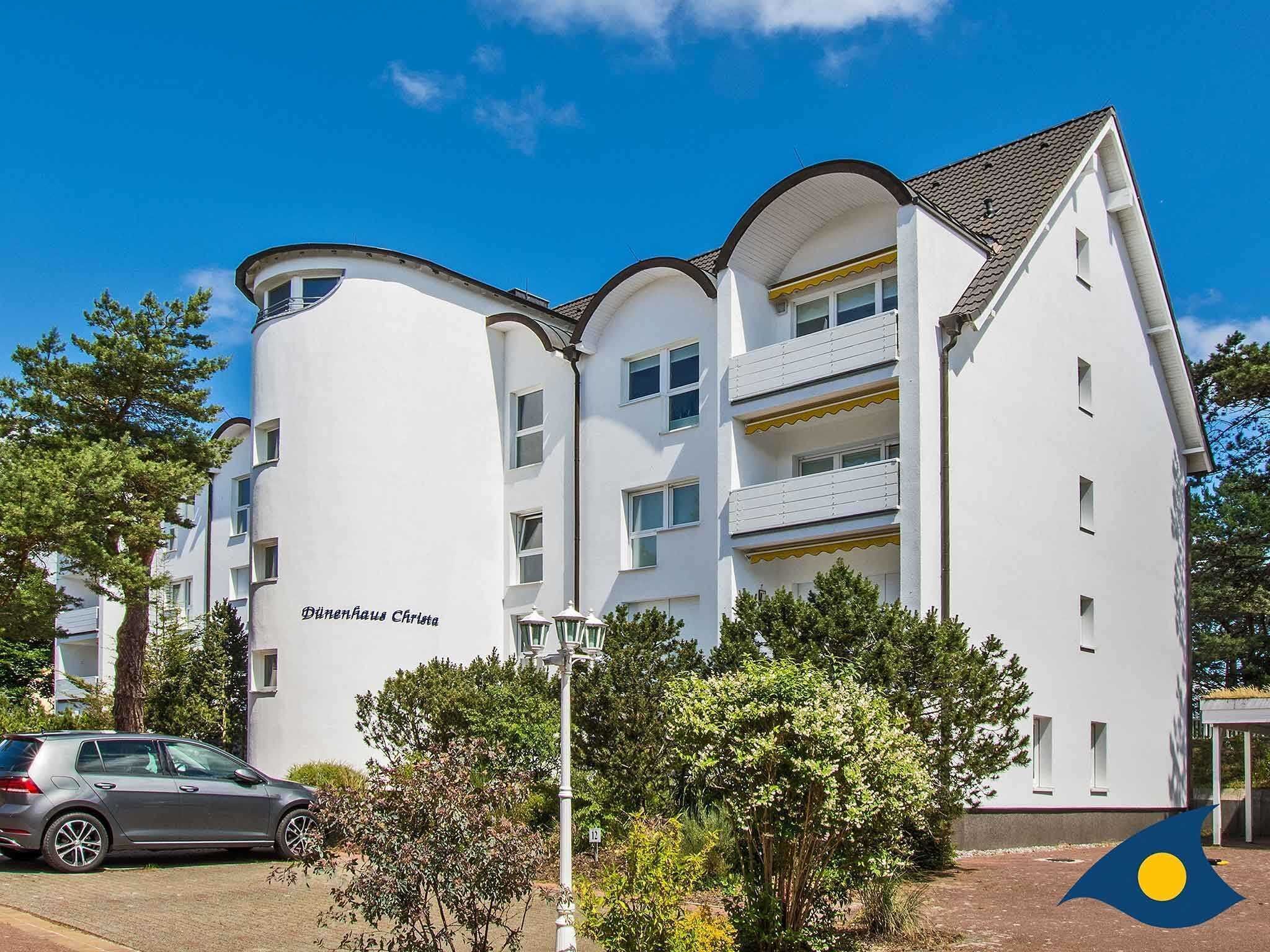 A multi-story building with a white facade, balconies, and a car parked in front.