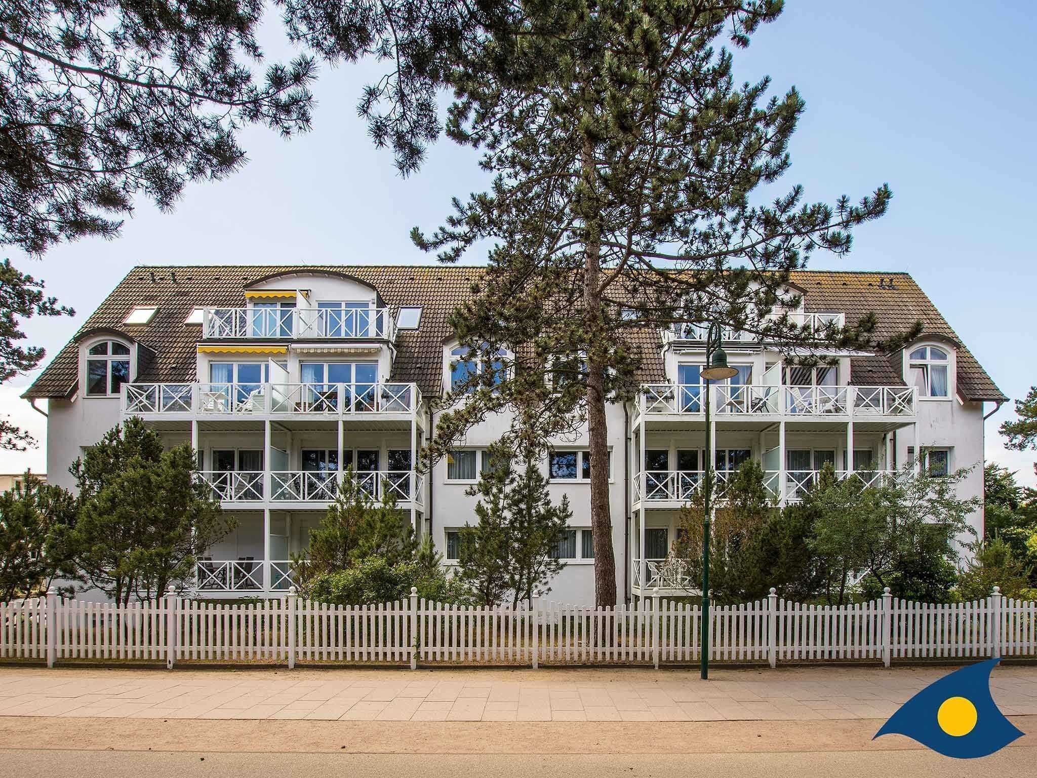 Un immeuble à plusieurs étages doté de balcons, d’une clôture blanche en bois et d’arbres au premier plan.