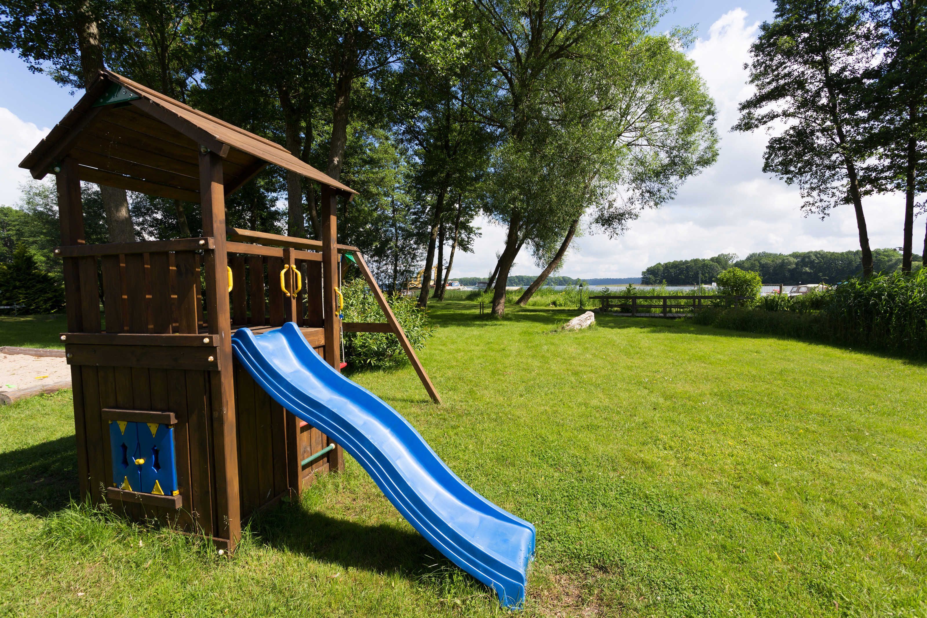 A wooden play structure with a blue slide in a grassy area with trees and a distant view of a body of water.