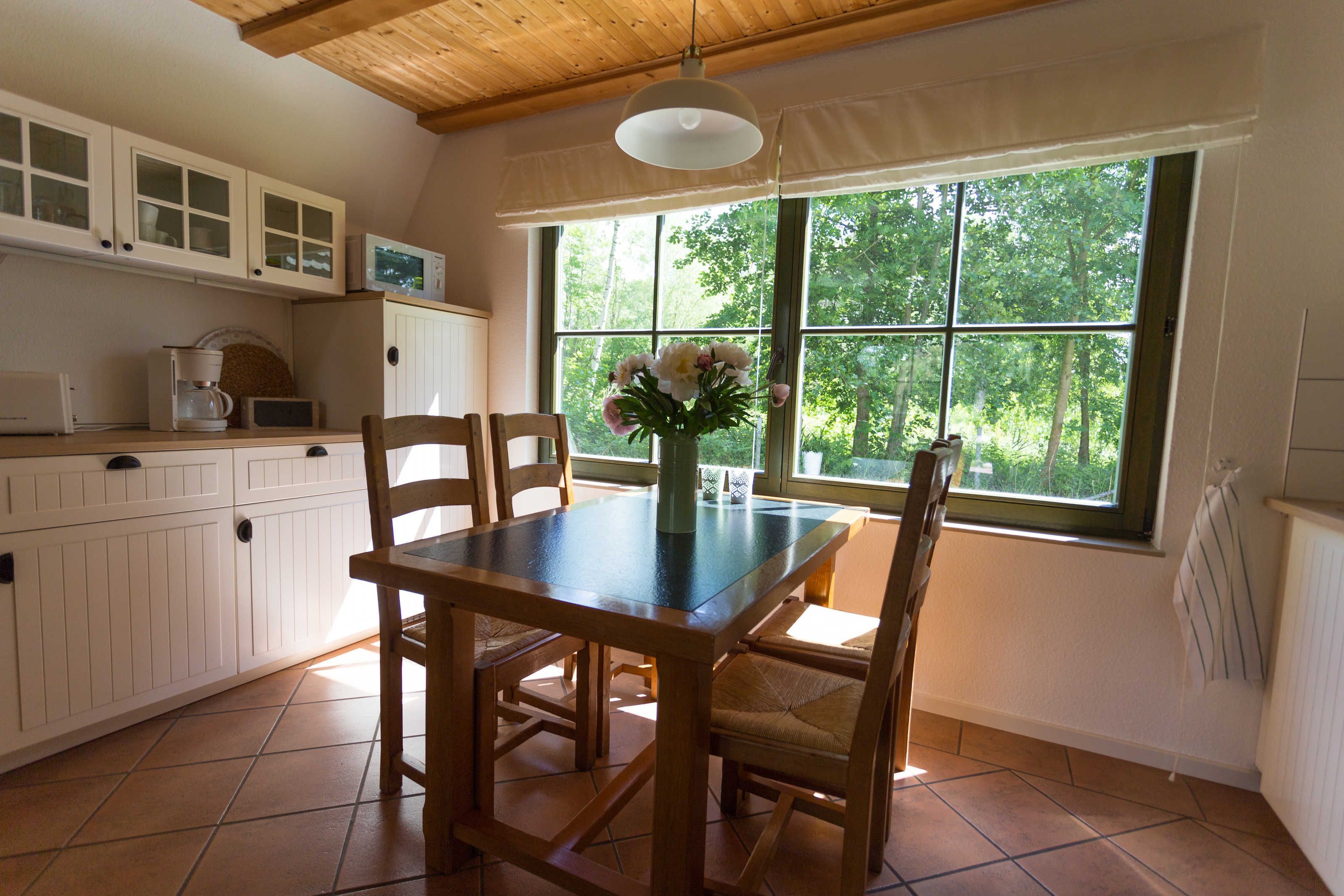 Une cuisine avec des meubles en bois, une table à manger avec des chaises, une fenêtre donnant sur les arbres et un luminaire suspendu.