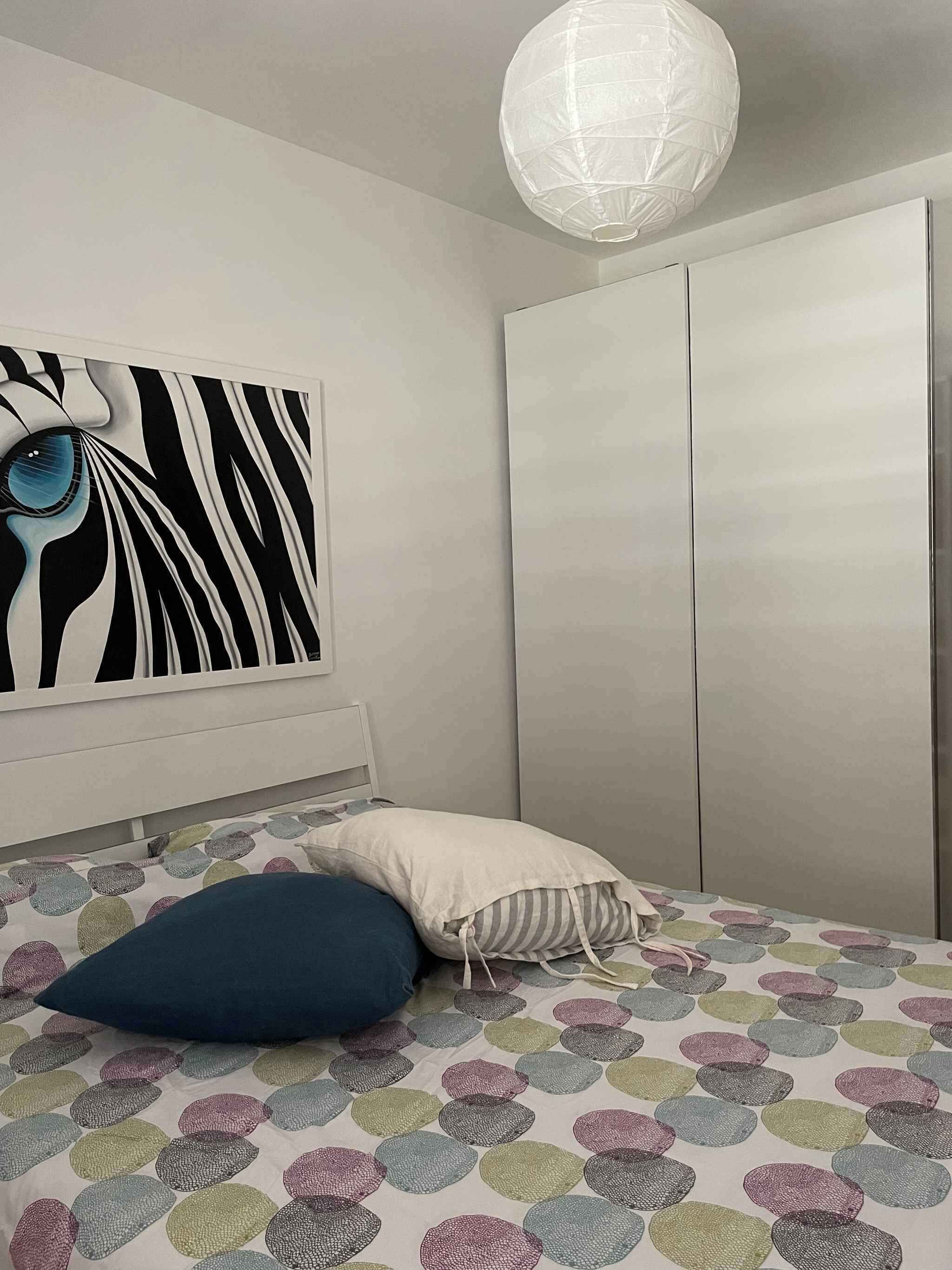 A bedroom with a bed, a zebra-striped wall art, a white wardrobe, and a hanging light fixture.