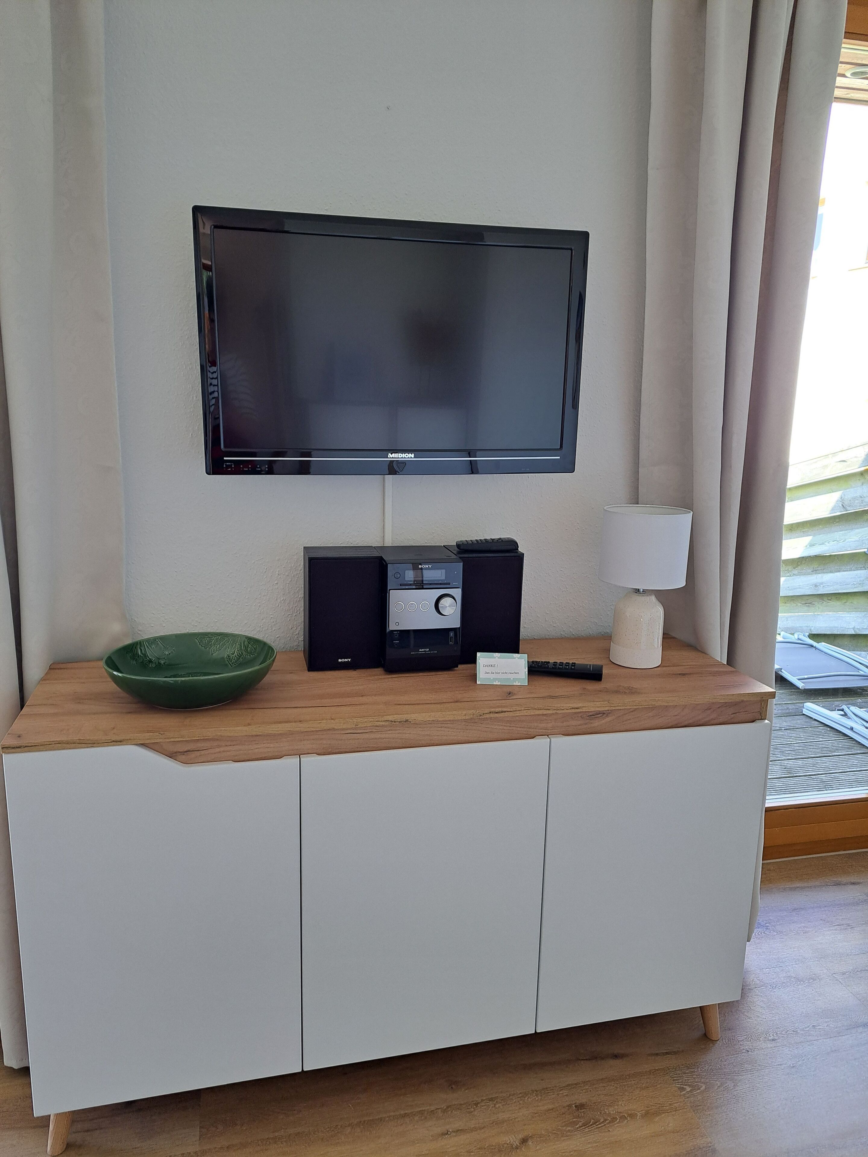 A flat-screen TV mounted on a wall, a wooden console table with a green bowl, a Sony audio system, a lamp, and a sign indicating a room number.