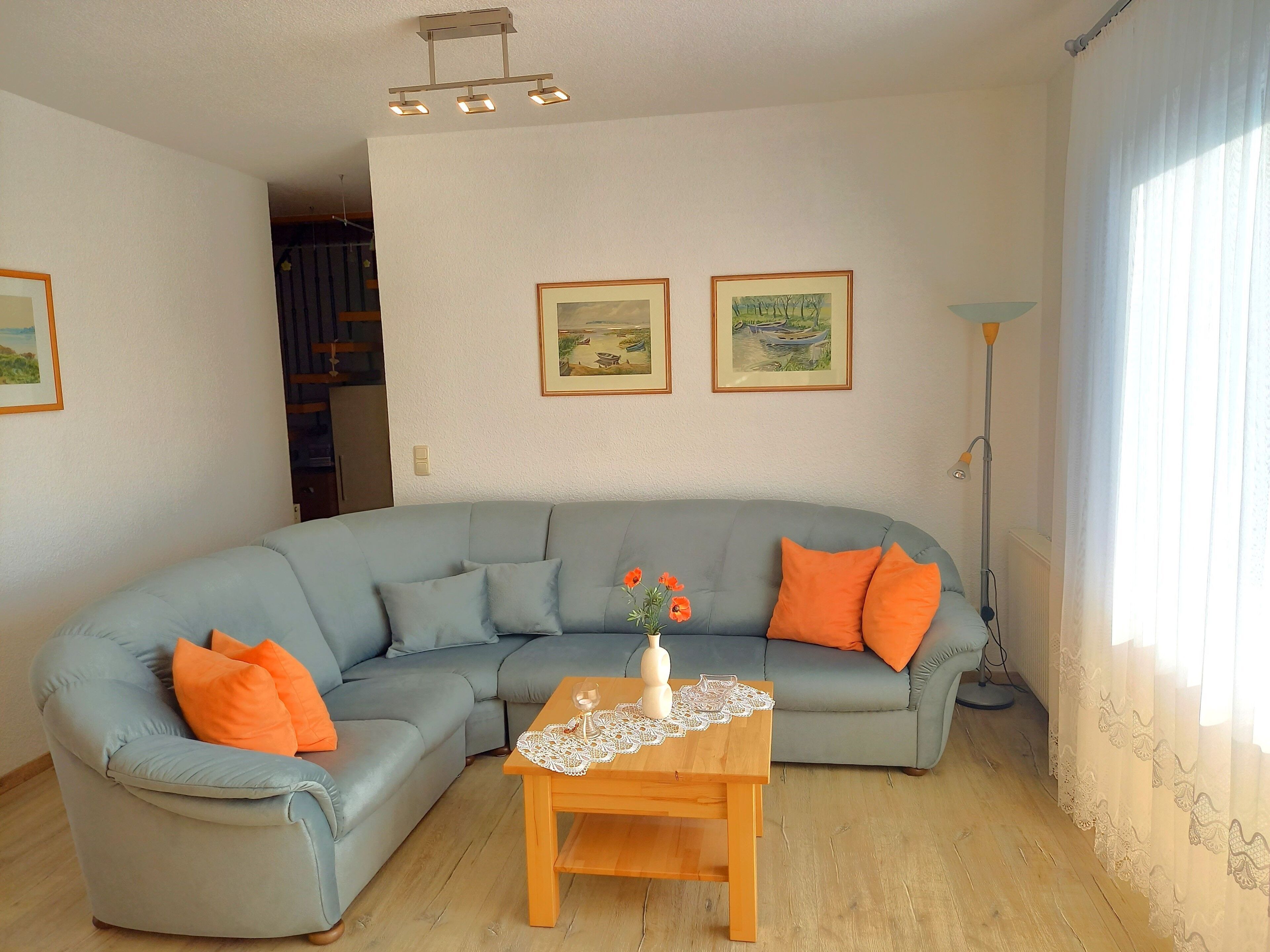 Un salón con un sofá gris, cojines anaranjados, una mesa de centro de madera con un posavasos de encaje, y dos cuadros de paisajes enmarcados en la pared.