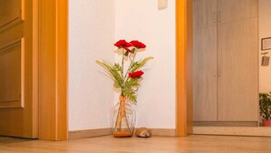 Um corredor com portas de madeira, um vaso de vidro com flores vermelhas e uma pequena estatueta de ouriço no chão.