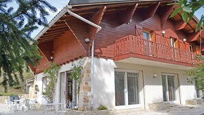 Una casa de madera con balcón, zócalo de piedra y un patio con mobiliario exterior.