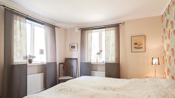 A bedroom with a large bed, two windows with curtains, a radiator, a chair, a picture on the wall, and a lamp. - 2 bedroom stunning home in Slite (Slite)