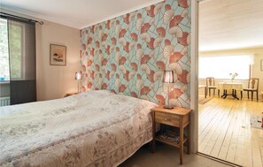 Une chambre à coucher comprenant un lit, une table de chevet, une lampe, un papier peint à motifs, un tableau encadré et une vue sur un coin repas, visible à travers une porte ouverte.