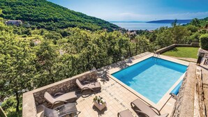 Una piscina rectangular con solárium de piedra y sillas de descanso, rodeada de exuberante vegetación y vistas a la costa distante.
