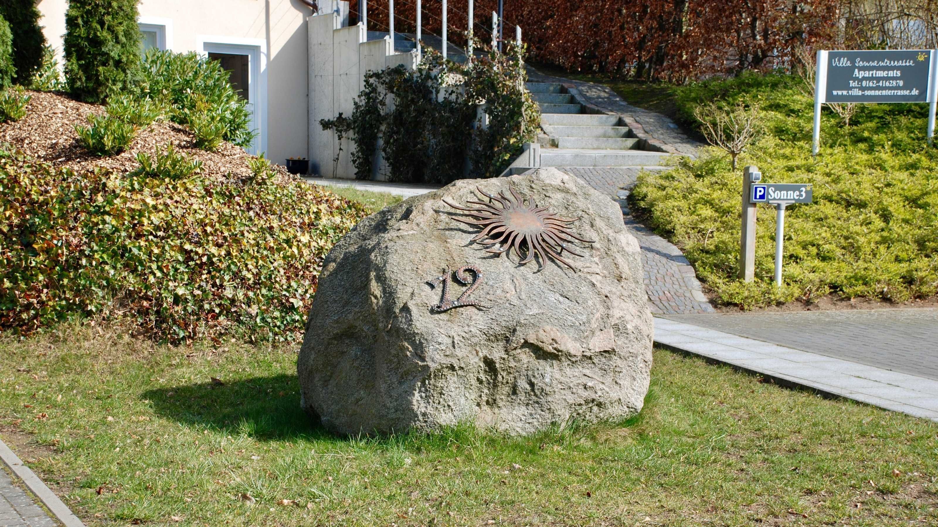 Ein großer Felsen mit Sonnendesign und der Zahl 12, ein Weg zu einem Gebäude und ein Schild für die Villa Sonnenterrasse Apartments.
