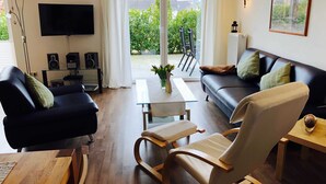 A modern living room with a black leather sofa, a wooden rocking chair, a glass coffee table, and a TV mounted on the wall. - Villa Sun 4, 2-5 P. Terrace - Villa Sun Terrace, Nicole Struck (Neustadt in Holstein)