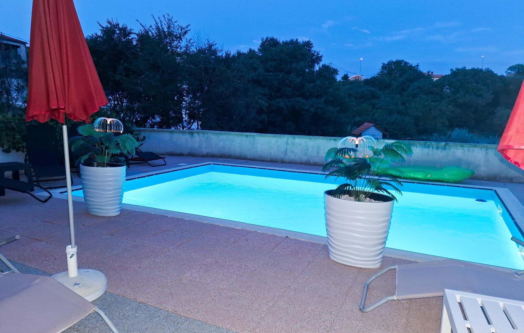 Una piscina con due piante in vaso, un ombrellone rosso e una sedia a sdraio.