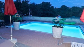 Une piscine agrémentée de deux plantes en pot, d’un parasol rouge et d’une chaise longue.