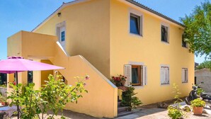 Une maison jaune à deux étages, avec un petit porche, un parasol rose et des plantes en pot.