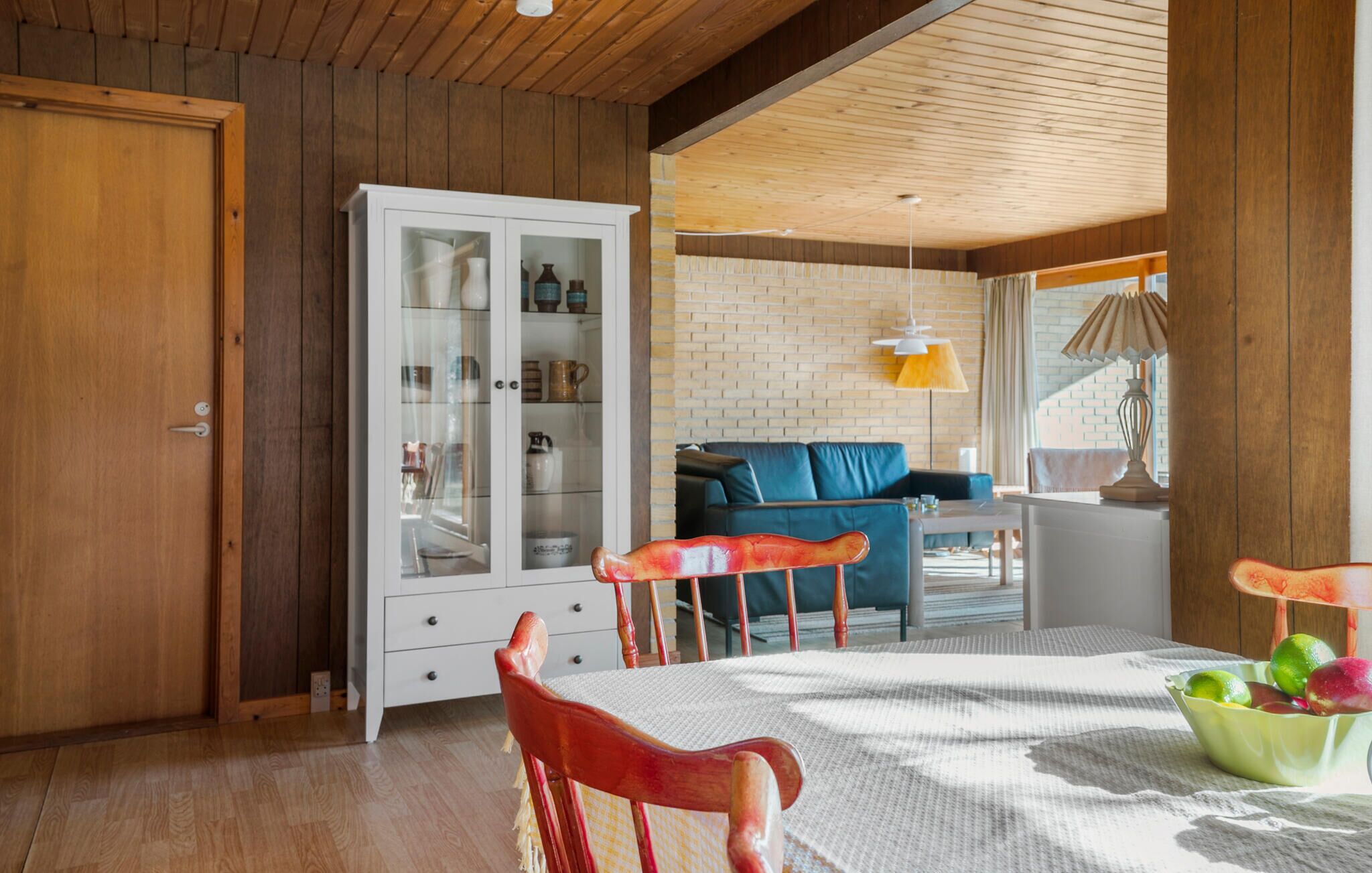 A cozy room with a wooden ceiling, a white cabinet with glass doors, a blue sofa, and a dining table with chairs.
