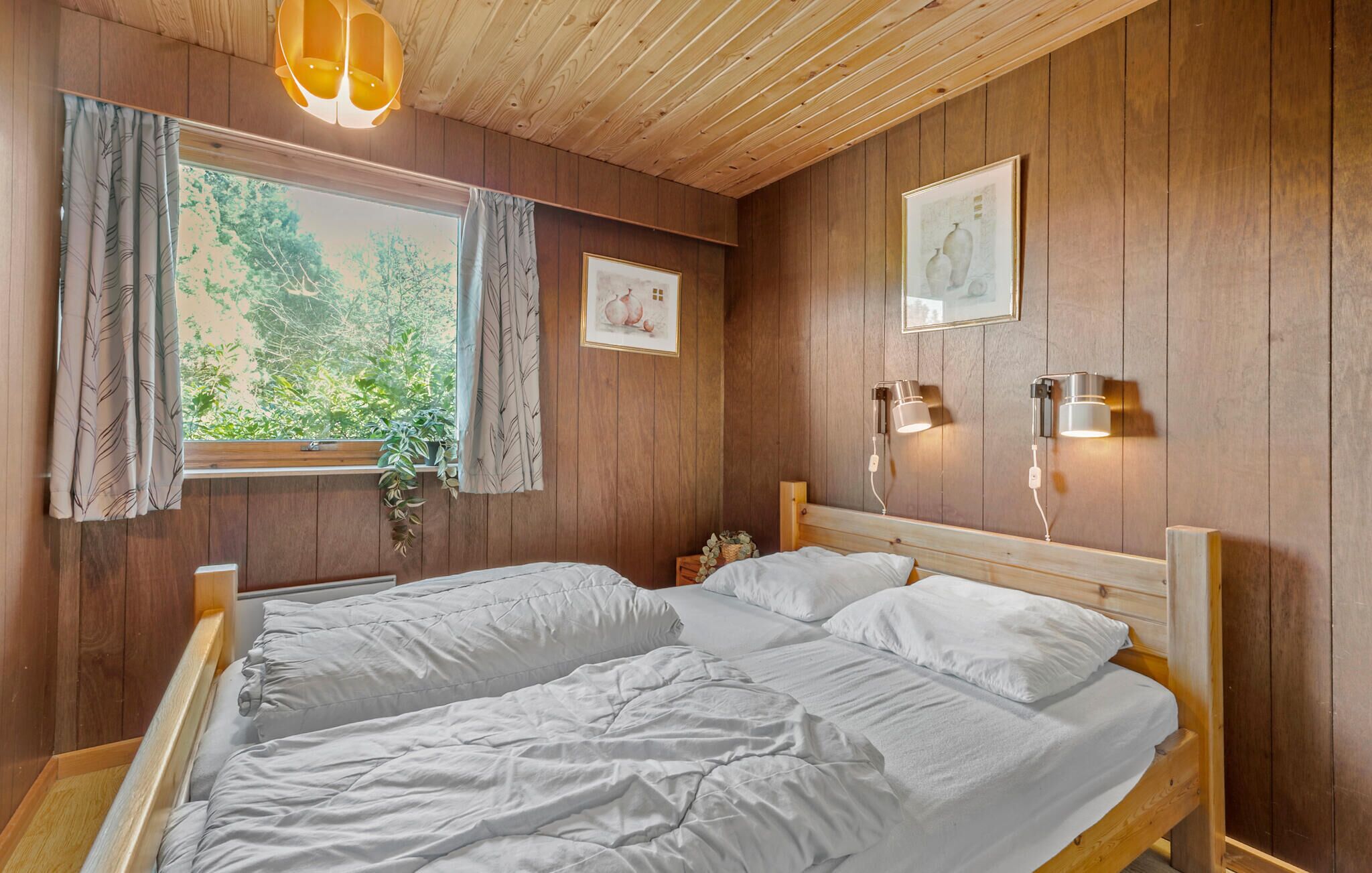 A wooden-paneled room with a double bed, two wall-mounted lamps, and a window with curtains.
