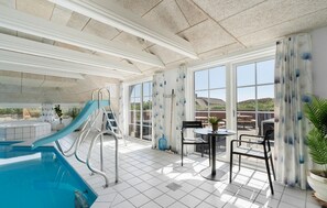 En indendørs swimmingpool med rutsjebane, solterrasse med bord og stole, samt store vinduer der giver udsigt til det fri.