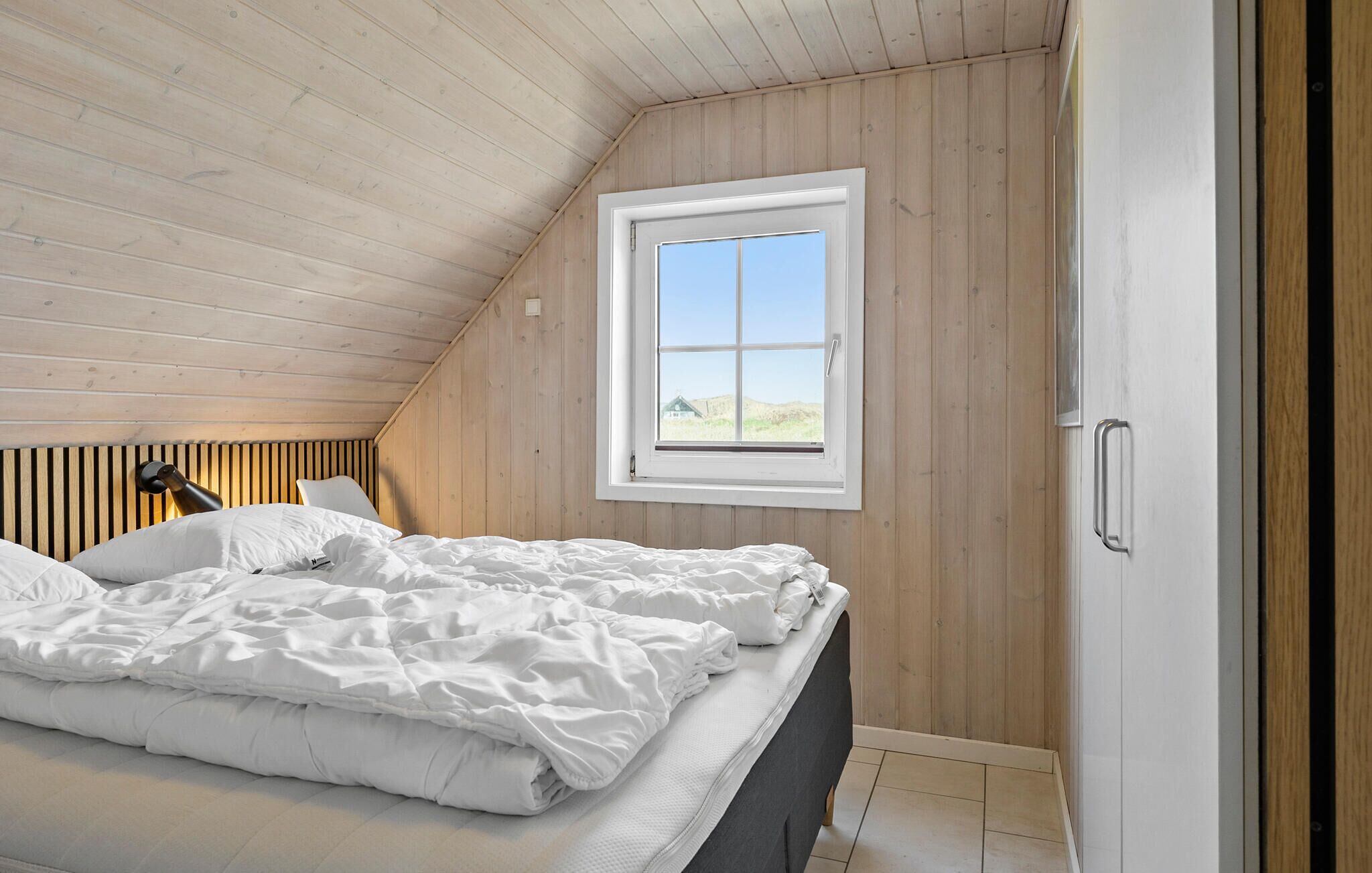 Une chambre avec un plafond en pente, une fenêtre donnant sur l’extérieur, un lit avec une literie blanche et une armoire en bois.