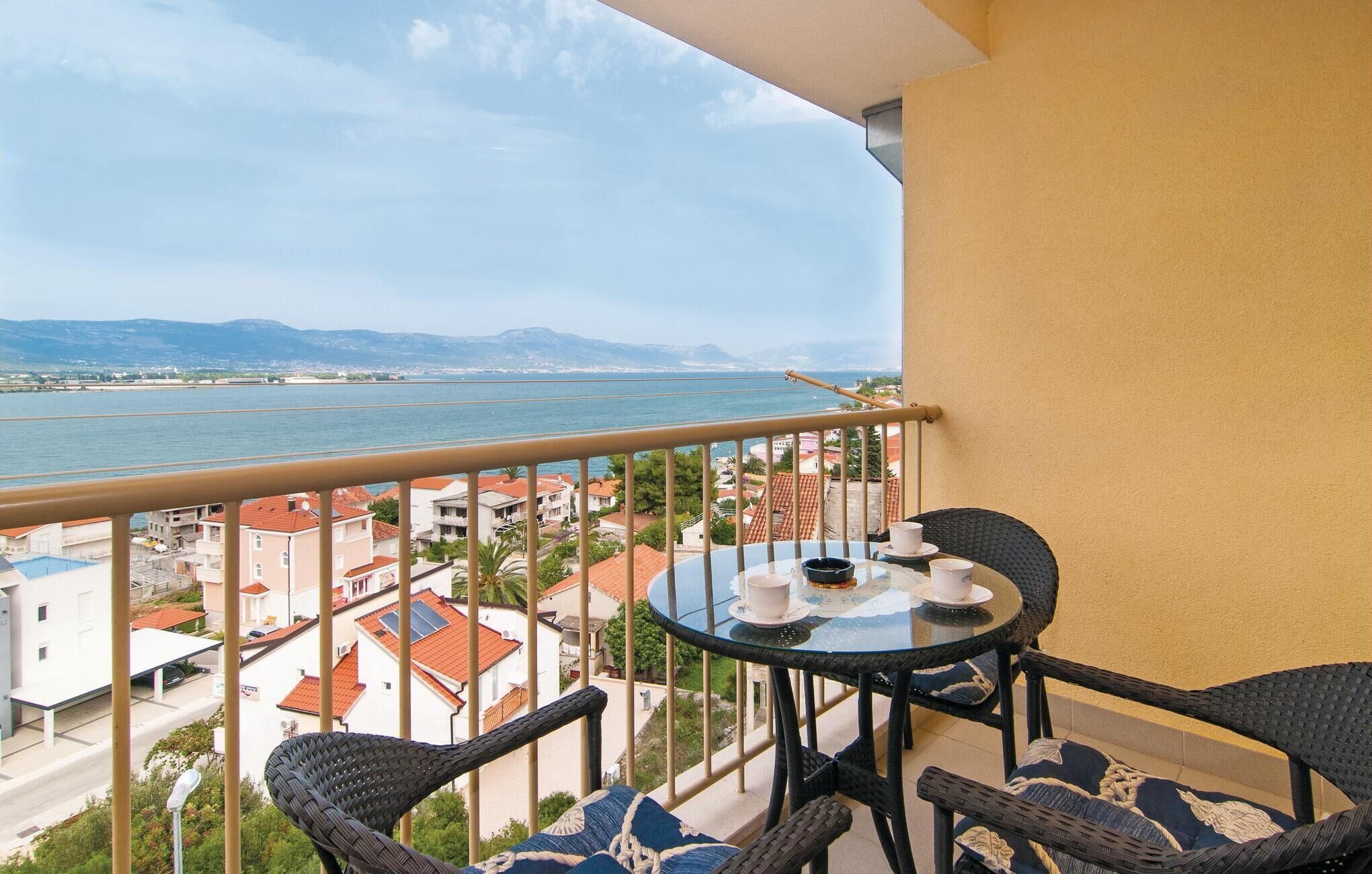 A balcony with a table and chairs, overlooking a coastal town and the sea.
