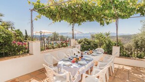 Un área de comedor al aire libre con una mesa preparada para cuatro personas, rodeada de sillas de plástico blanco, bajo una pérgola con parras.