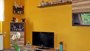 Una sala de estar con televisor, estantes con figuritas, una planta en maceta y una ventana.