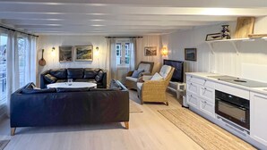 Ett modernt vardagsrum med en svart soffa, ett runt soffbord och en fåtölj i rotting. Här finns också en köksdel med vita skåp och en spis.
