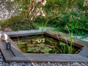 Eine Holzterrasse umgibt einen rechteckigen Teich mit Seerosen und einer Laterne.
