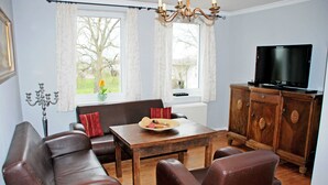 Ein Wohnzimmer mit einer Couch, Sesseln, einem Tisch mit einem Teller Essen, einem Fernseher und einem Schrank.