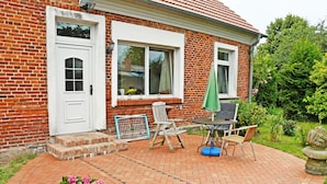 Ein Backsteinhaus mit weißer Tür und Fenstern, eine Terrasse mit Tisch und Stühlen und ein Garten mit Blumen.