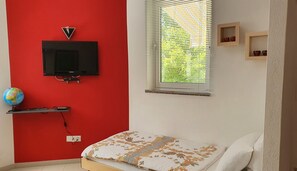 Een slaapkamer met een rode wand, een televisie bevestigd boven een plank, een wereldbol en een bed met een geborduurde spreideken.