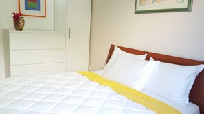 Un lit avec une literie blanche et un détail jaune, une commode blanche et un tableau encadré au mur.