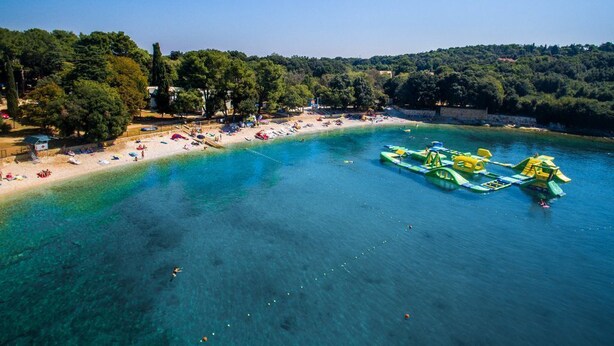 Vue aérienne d’une plage aux eaux turquoise et limpides, bordée d’un rivage sablonneux et ponctuée de quelques embarcations vertes.