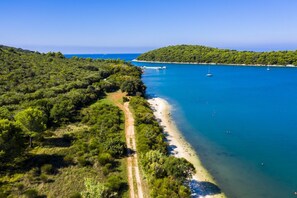 A coastal landscape with a dirt path leading to a beach, boats on the water, and a forested hill in the background. - Ginkgo 3 - Apartment near the sea - Apartments Ginkgo, Pula-Banjole (Banjole)