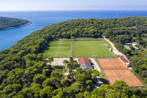 Une vue aérienne d’un complexe sportif comprenant plusieurs courts de tennis, entouré d’une forêt dense et avec la côte en arrière-plan.