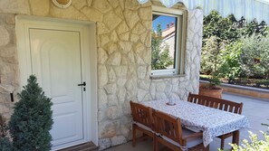 Un bâtiment en pierre avec une porte et une fenêtre blanches, une petite table recouverte d’une nappe à motifs, des chaises en bois et une plante en pot.