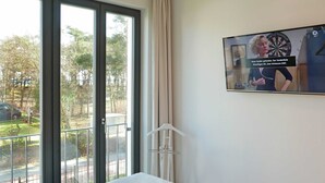 A hotel room with a bed, a television displaying a dartboard, a window with a view of trees and a road, and a white hanger on the wall.