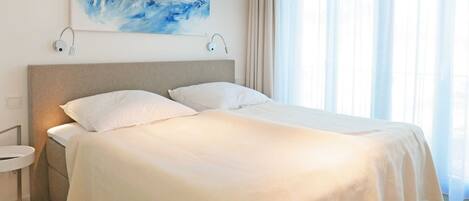 Una cama bien tendida con ropa de cama blanca en una habitación de diseño moderno, un cuadro en la pared y una ventana con cortinas translúcidas.