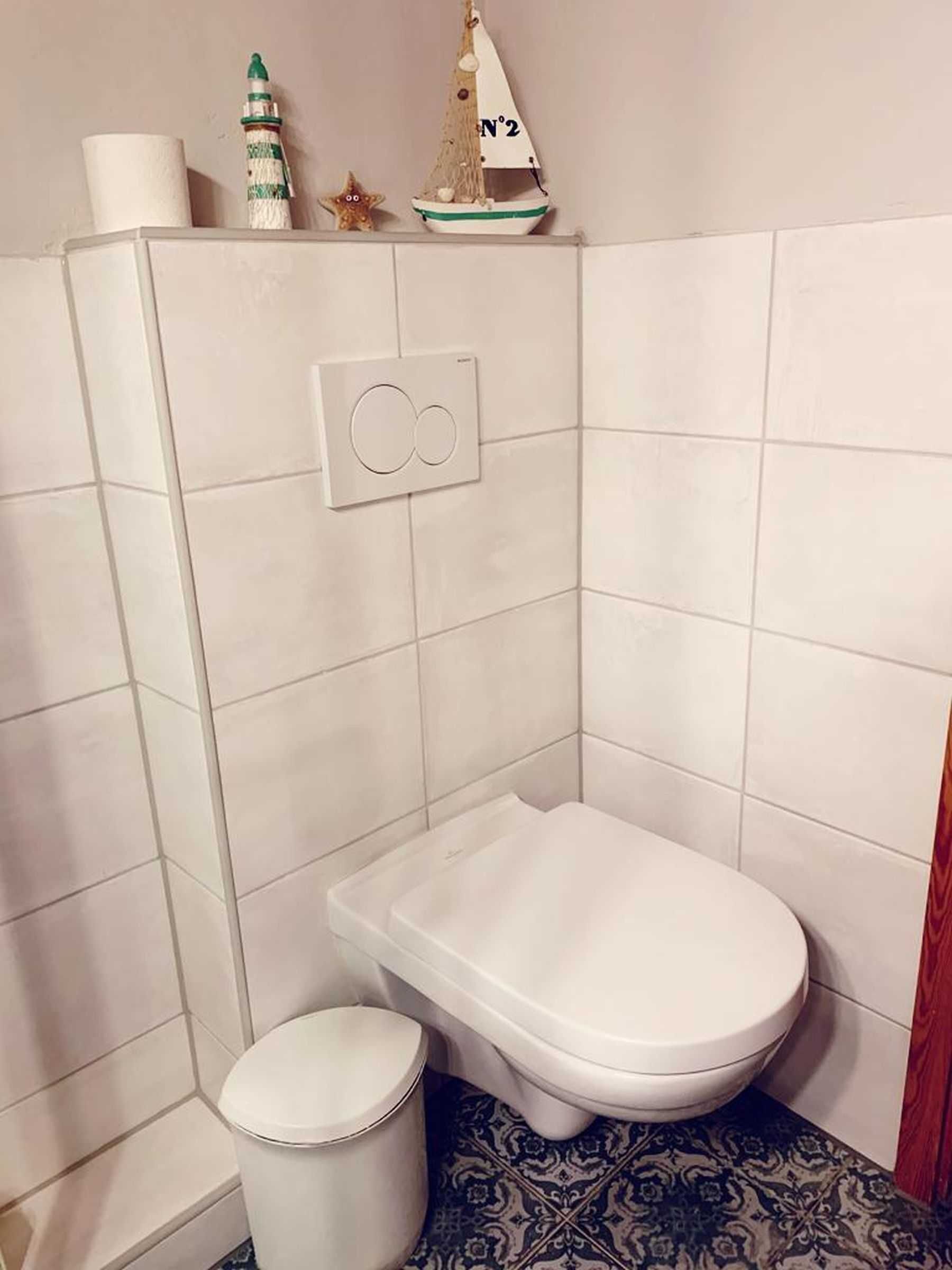 Et lille, kompakt badeværelse med et hvidt toilet, en skraldespand og en dekorativ sejlbåd på væggen.
