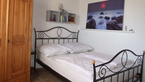 A bedroom with a bed, a wooden wardrobe, a shelf with books and decorative items, and a wall-mounted painting of a night scene.