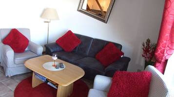 Un salon avec un canapé à carreaux, un canapé foncé et une table basse en bois. On trouve des coussins rouges sur les canapés et une lampe sur le sol.