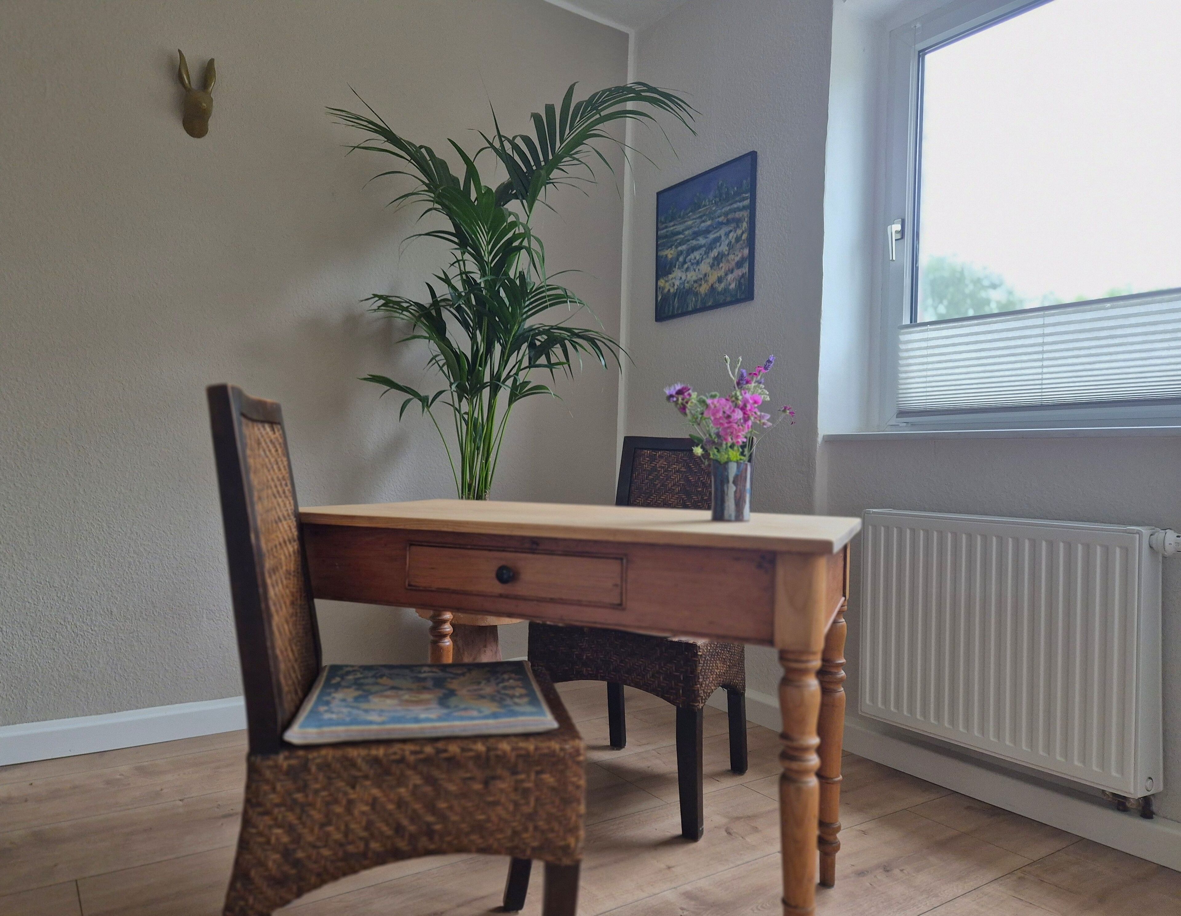 A dining area with a wooden table, two chairs, a potted plant, and a window with blinds.