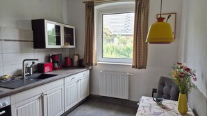 Een keuken met witte kastjes, een raam met gordijnen, een radiator en een tafel met een tafelkleed met bloemen.